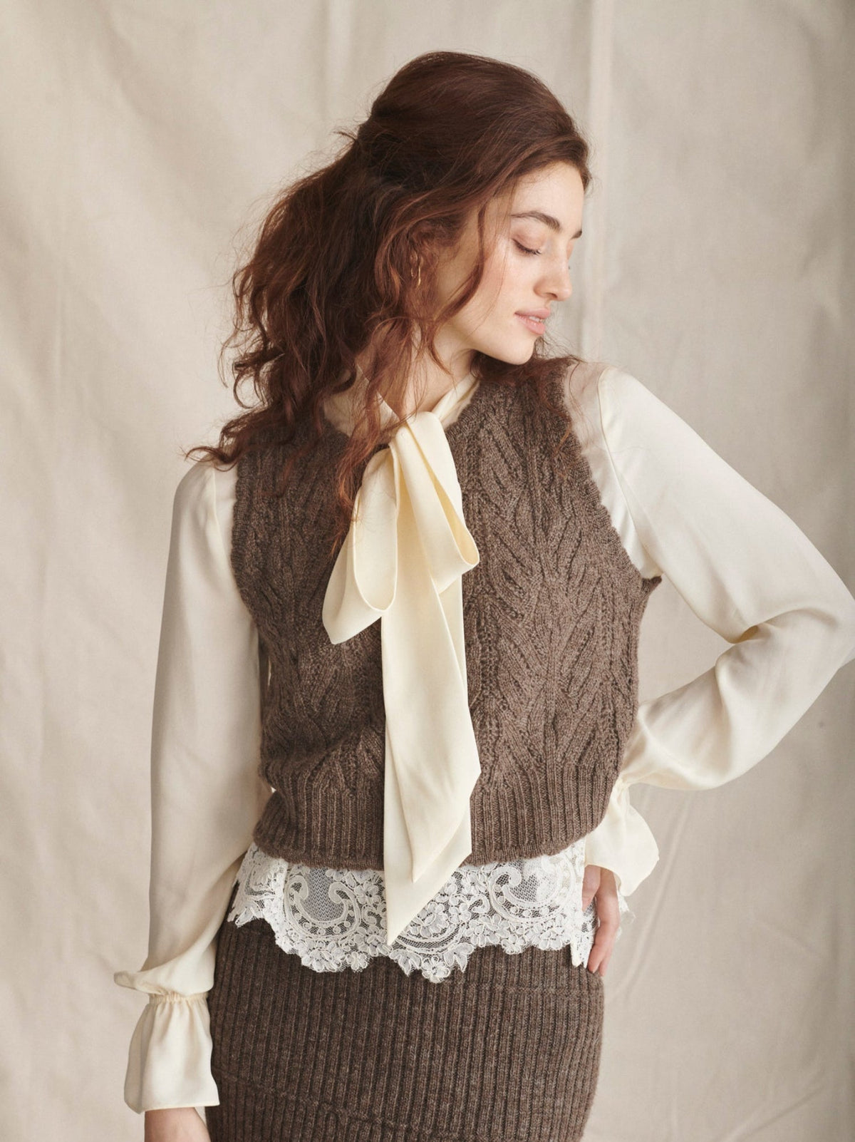 Woman wearing a brown knitted vest over a white blouse with a beige scarf, against a plain background.