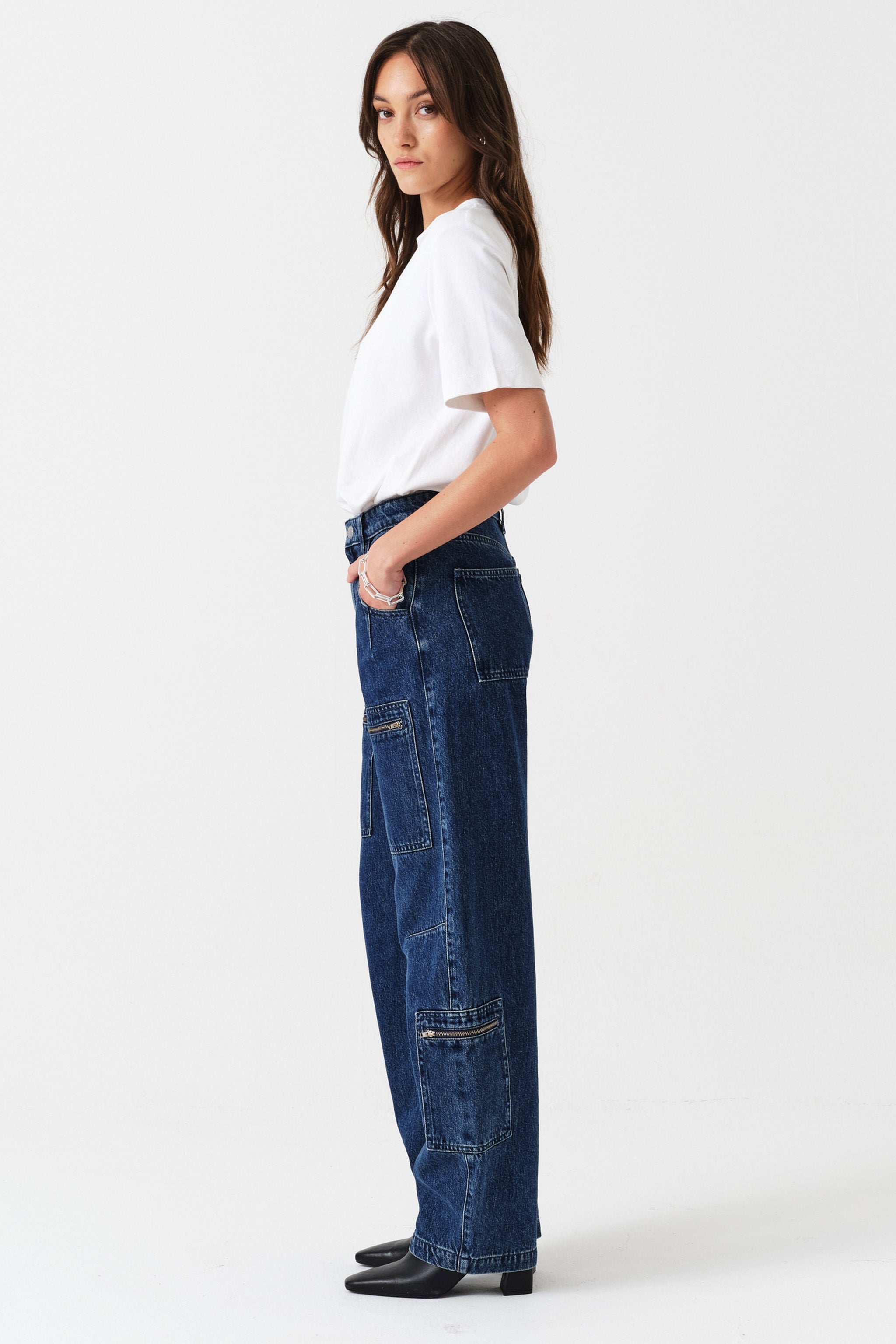Woman wearing a white t-shirt and blue jeans with zip details on a white background photographed from the side