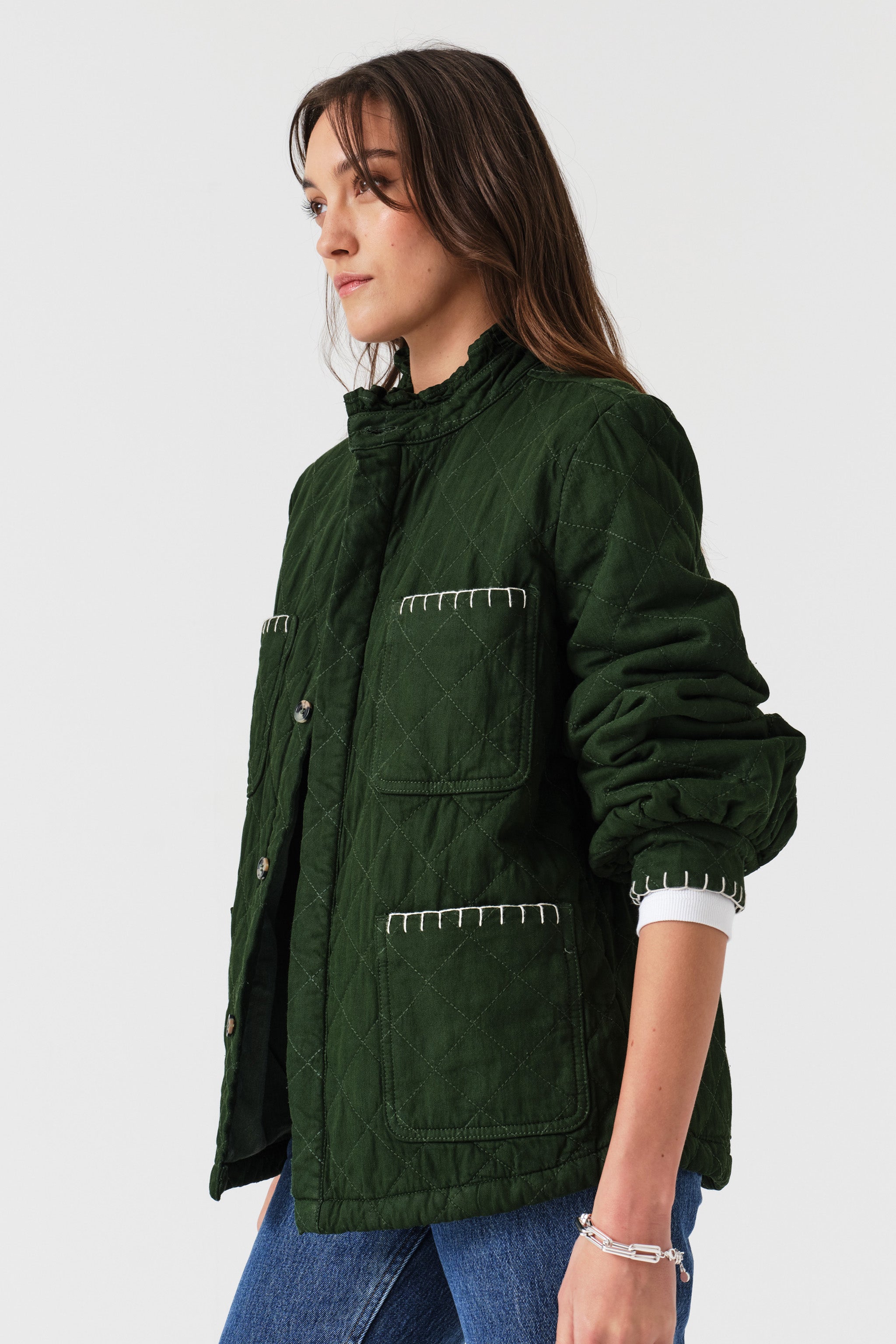 Woman wearing a quilted dark green jacket with white embroidered details and blue jeans on a white background