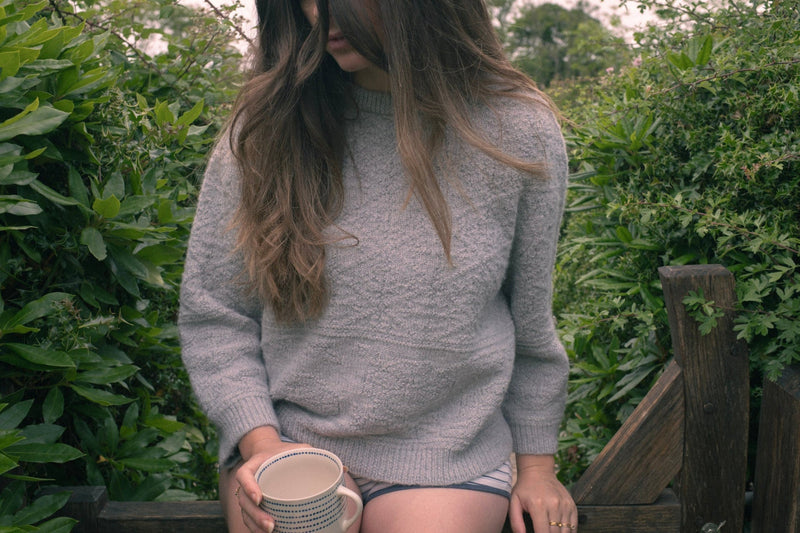 A woman wearing a Grey Cloud Stitch Jumper, showcasing textured detail in a natural outdoor setting. Sustainable knitwear jumper.