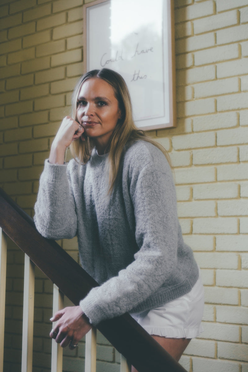 Woman in a gray sweater standing against a brick wall.