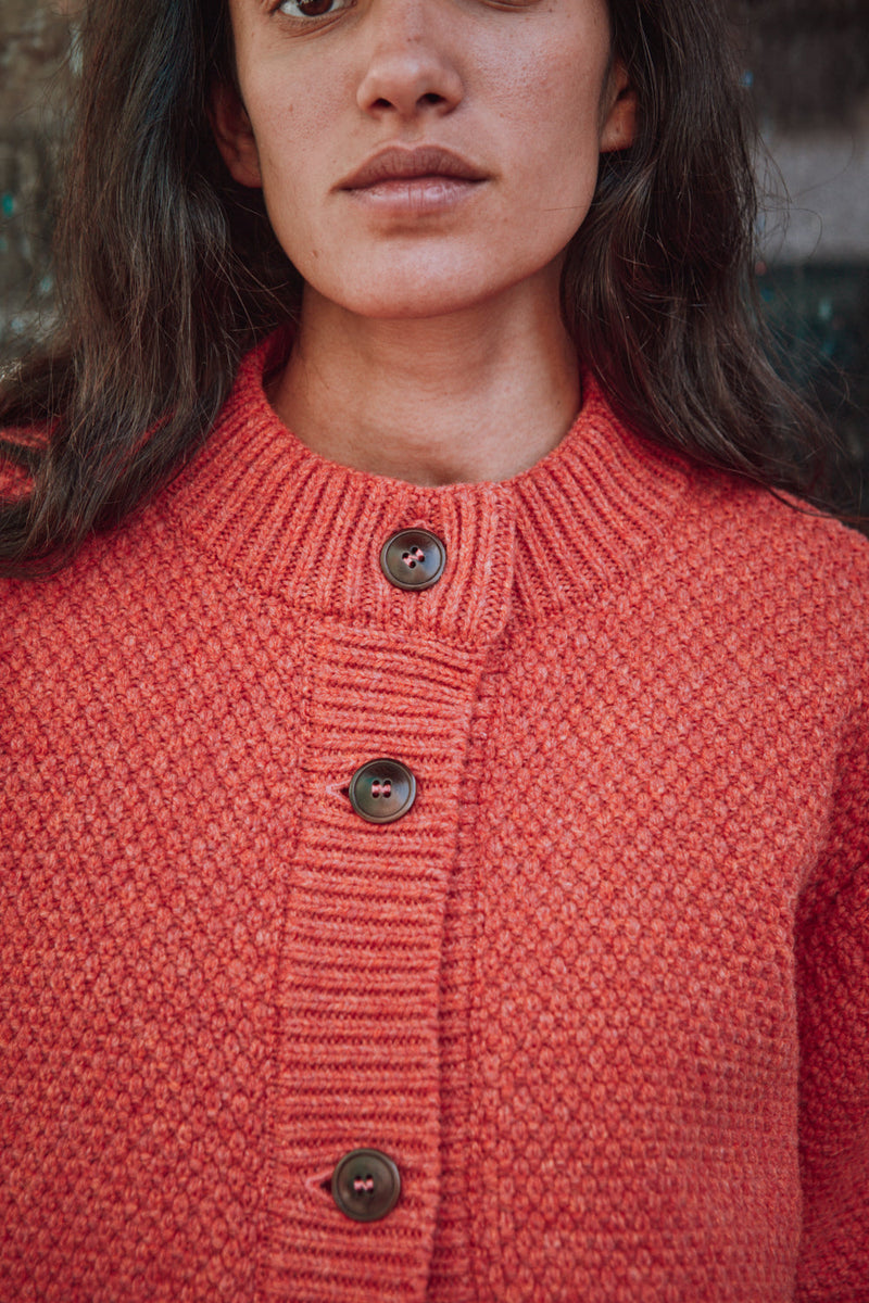 Person wearing a red knitted L'ENVERS cardigan standing in front of a textured wall.