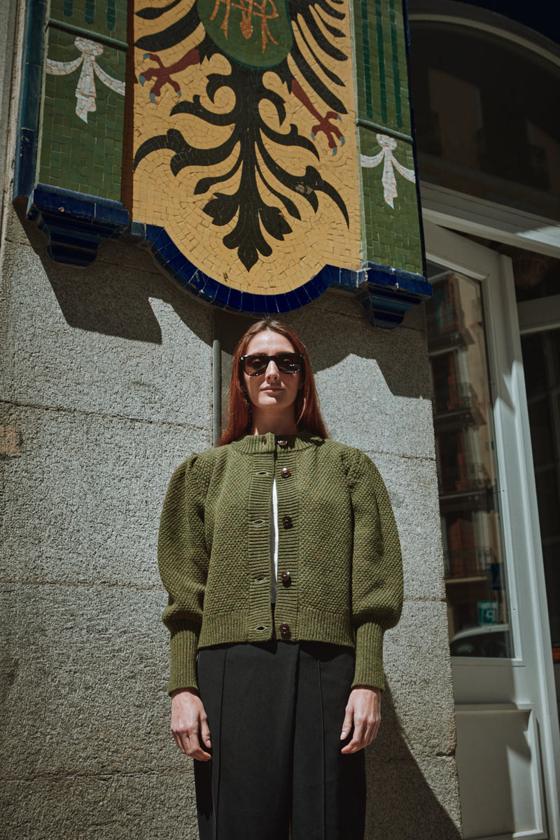 Woman wearing a green L'ENVERS cardigan standing in front of a building with a decorative emblem.