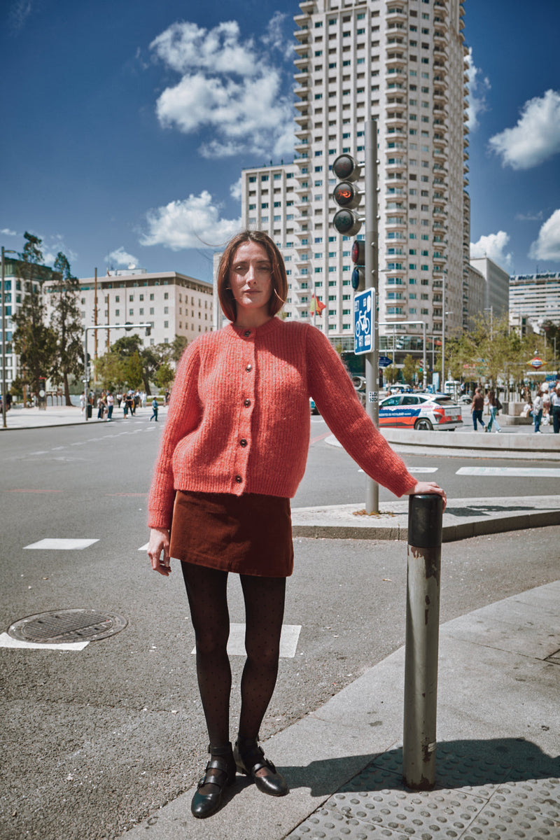 Woman in a pink sweater and skirt standing on a city street with tall buildings in the background.