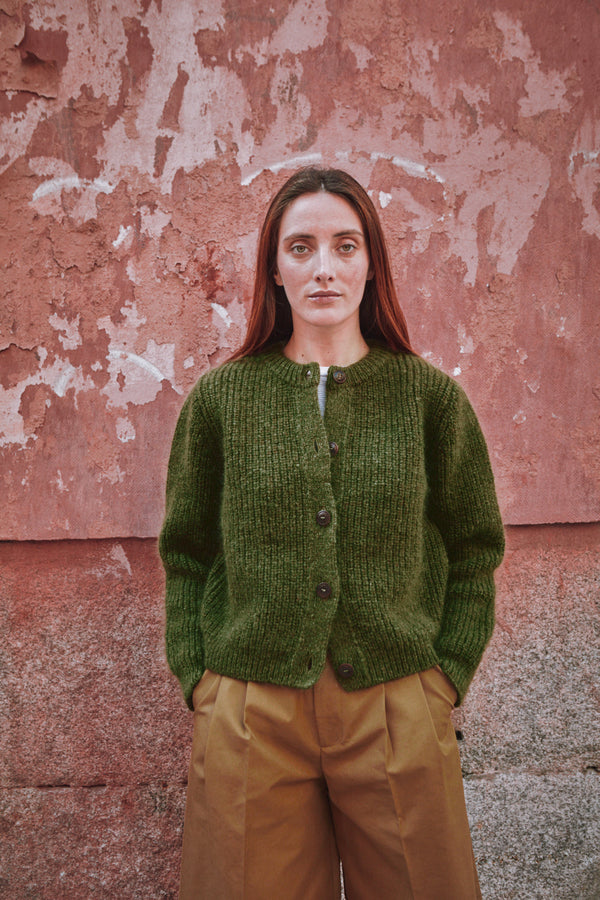 Woman wearing a green knitted cardigan against a textured pink wall.