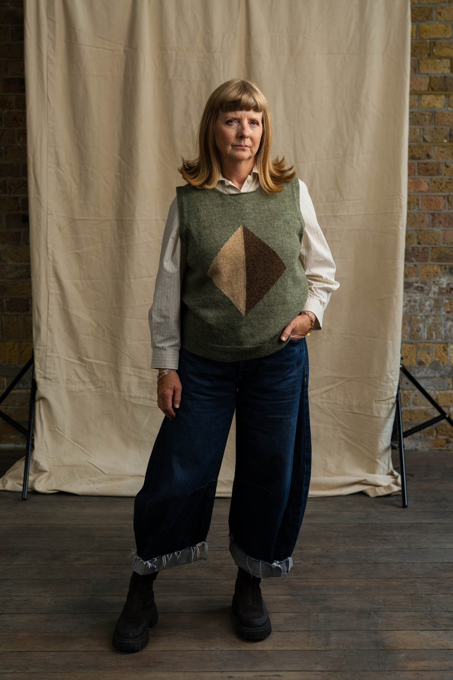 Woman wearing a patterned vest over a shirt, standing against a brick wall with a beige curtain.