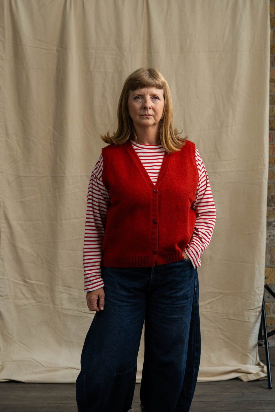 Person wearing a red vest over a striped shirt with blue jeans, standing against a beige backdrop.