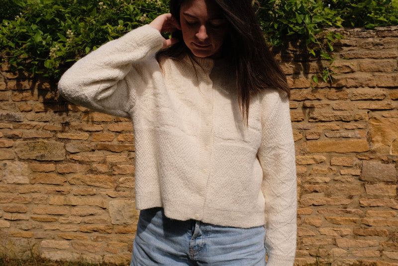 A woman wearing a relaxed fit ivory cardigan made from sustainable knitwear cardigan against a stone wall.