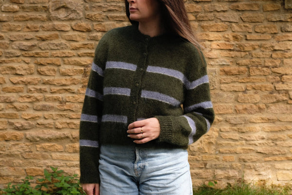 Person wearing a green and blue striped sweater with denim shorts standing against a stone wall.