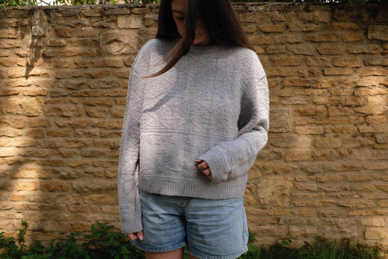 Model wearing a light grey sustainable knitwear jumper paired with denim shorts against a stone wall backdrop.