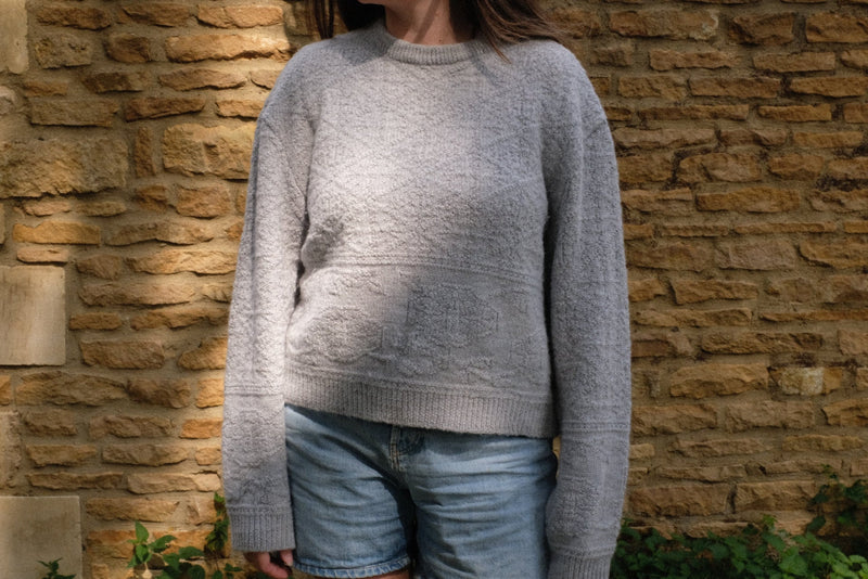Woman wearing a light grey sustainable knitwear jumper with textured detail, paired with denim shorts.