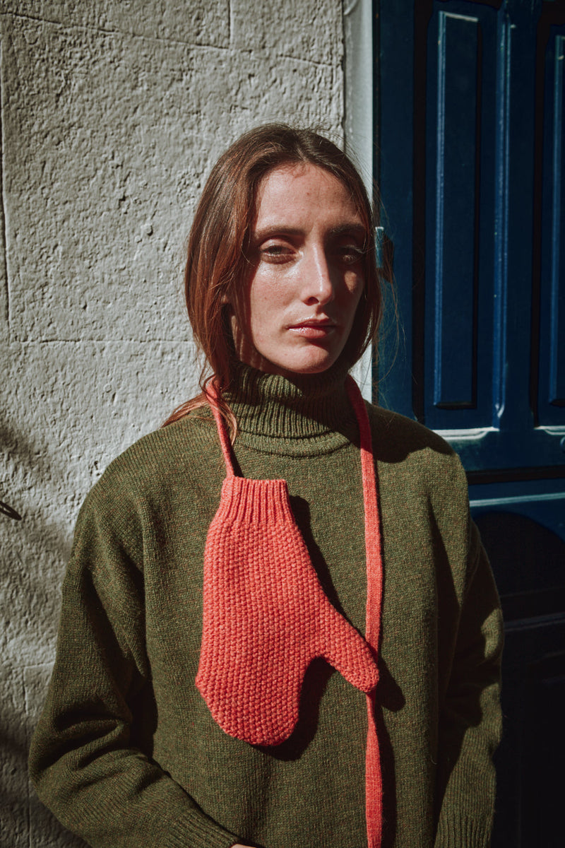 Woman wearing a green sweater with coral knitted mittens against a textured wall and blue door.