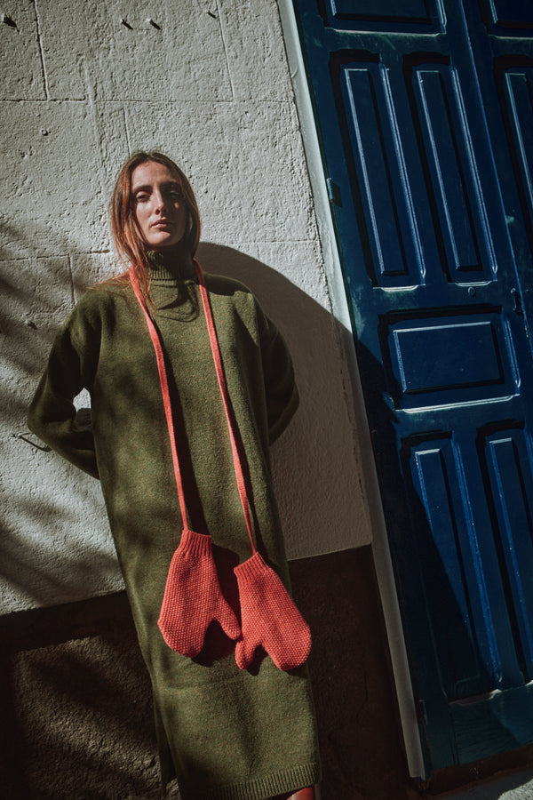 Woman wearing a green sweater with coral knitted mittens against a textured wall and blue door.