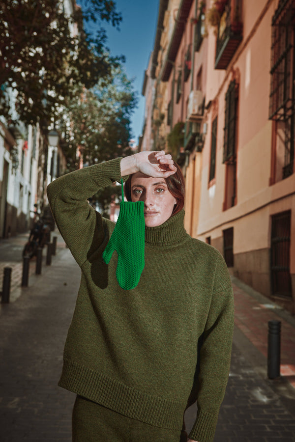 Person wearing a green sweater and green mittens on a street with buildings and trees in the background