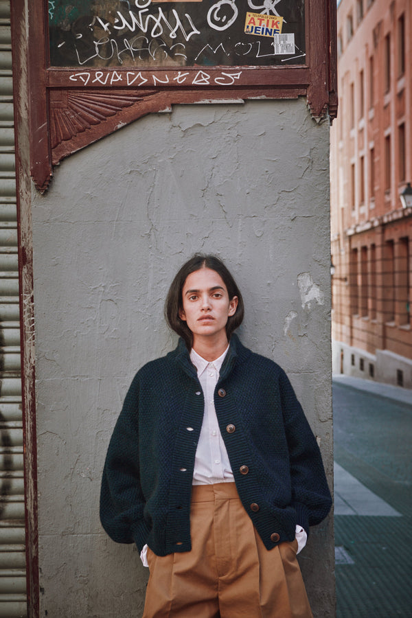 Person wearing a dark jacket and beige pants standing against a textured wall with graffiti.