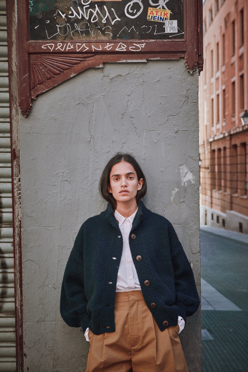 Person wearing a dark jacket and beige pants standing against a textured wall with graffiti.