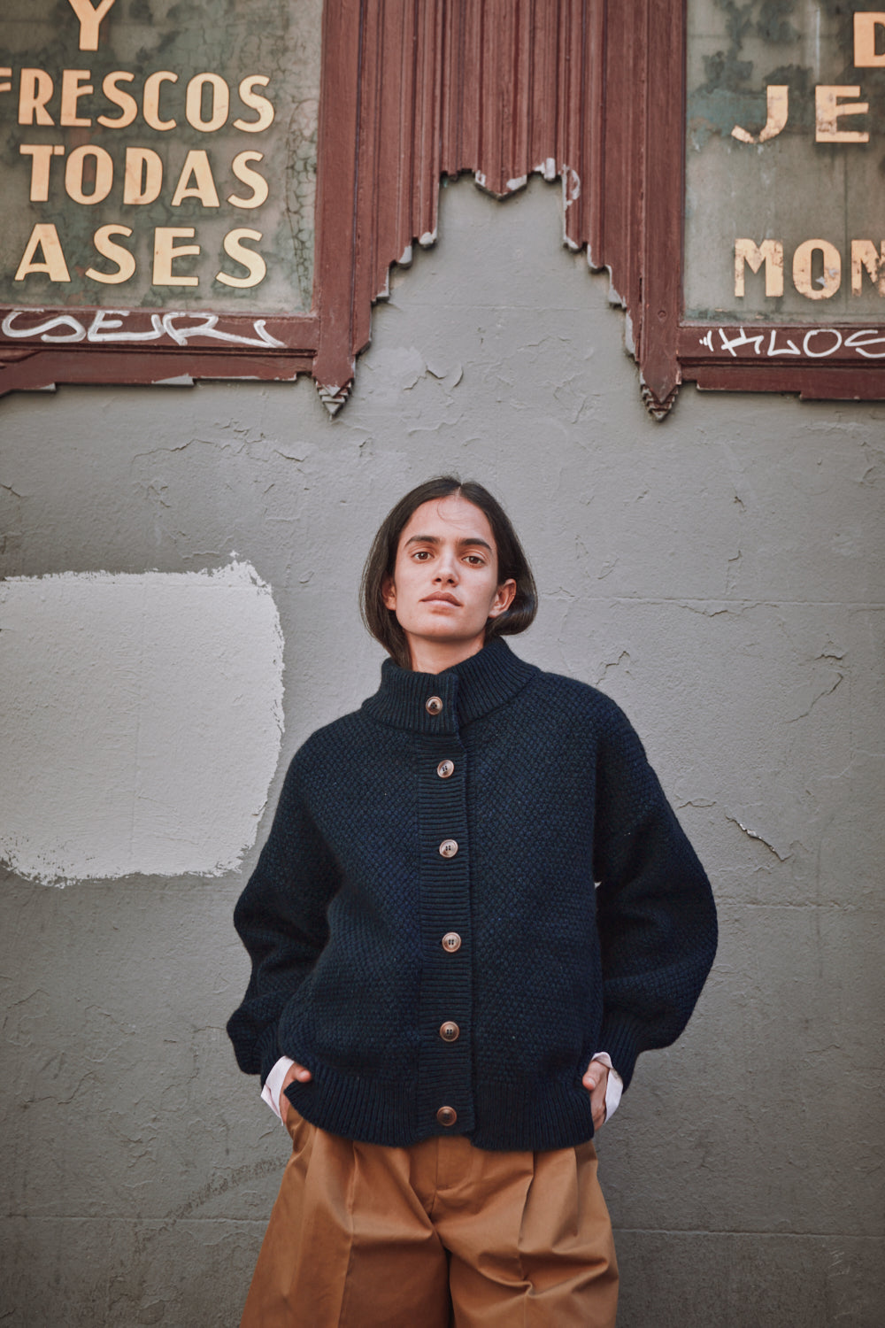 Person wearing a dark blue jacket with buttons against a textured wall with peeling paint and graffiti.