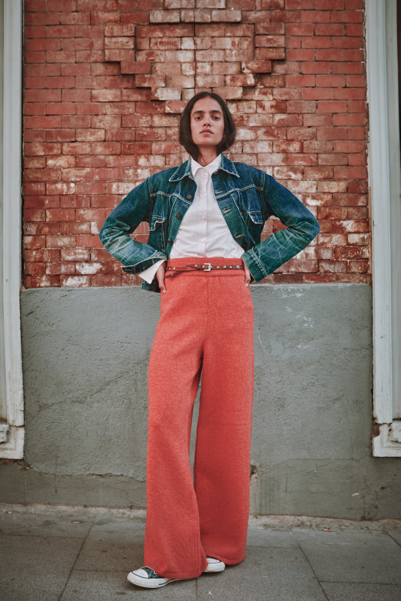 Person wearing a denim jacket, white shirt, and red pants standing against a brick wall.