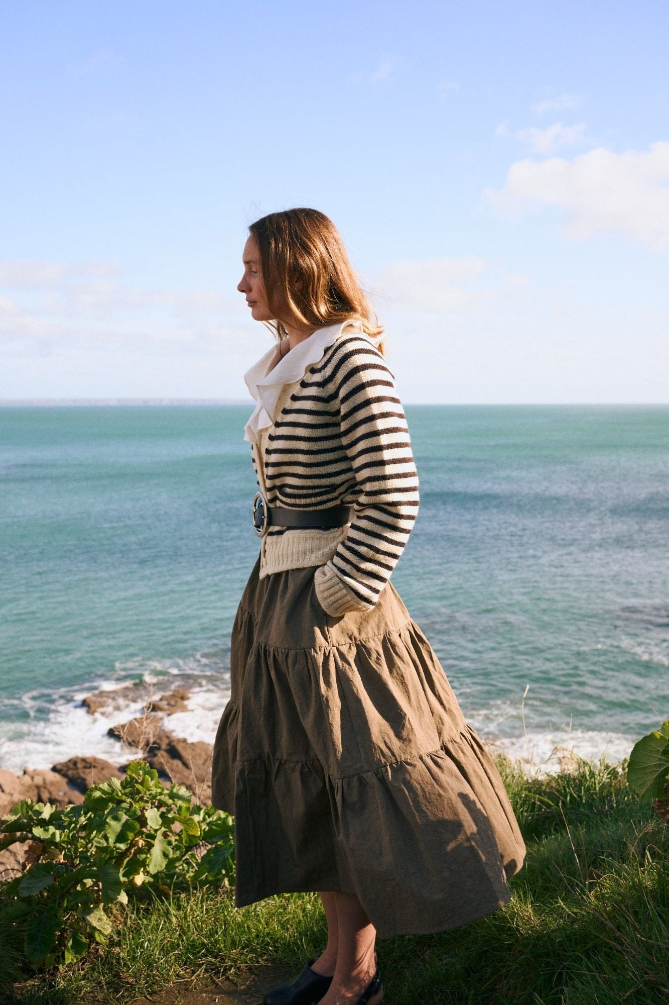 Woman standing on a cliff overlooking the ocean with a blue sky in a HERD cardigan in ecru and dark chocolate stripe. 