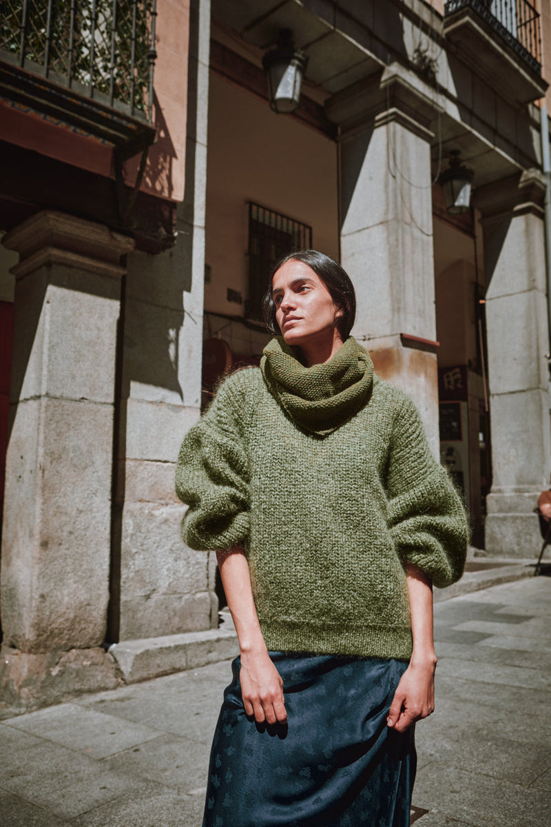 Person wearing a green sweater standing on a street with stone buildings.