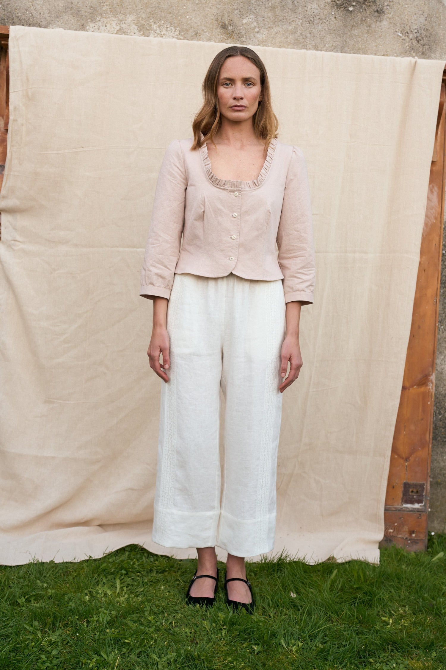 Woman wearing a beige top and white pants standing in front of a large beige fabric backdrop.