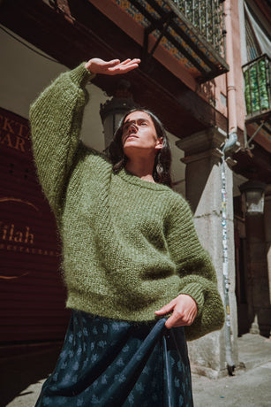 Woman wearing a green L'ENVERS sweater standing against a textured wall.