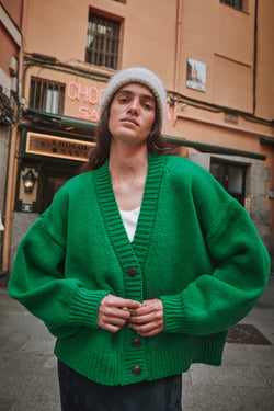 Woman wearing a green cardigan and white beanie standing on a street with a building in the background.
