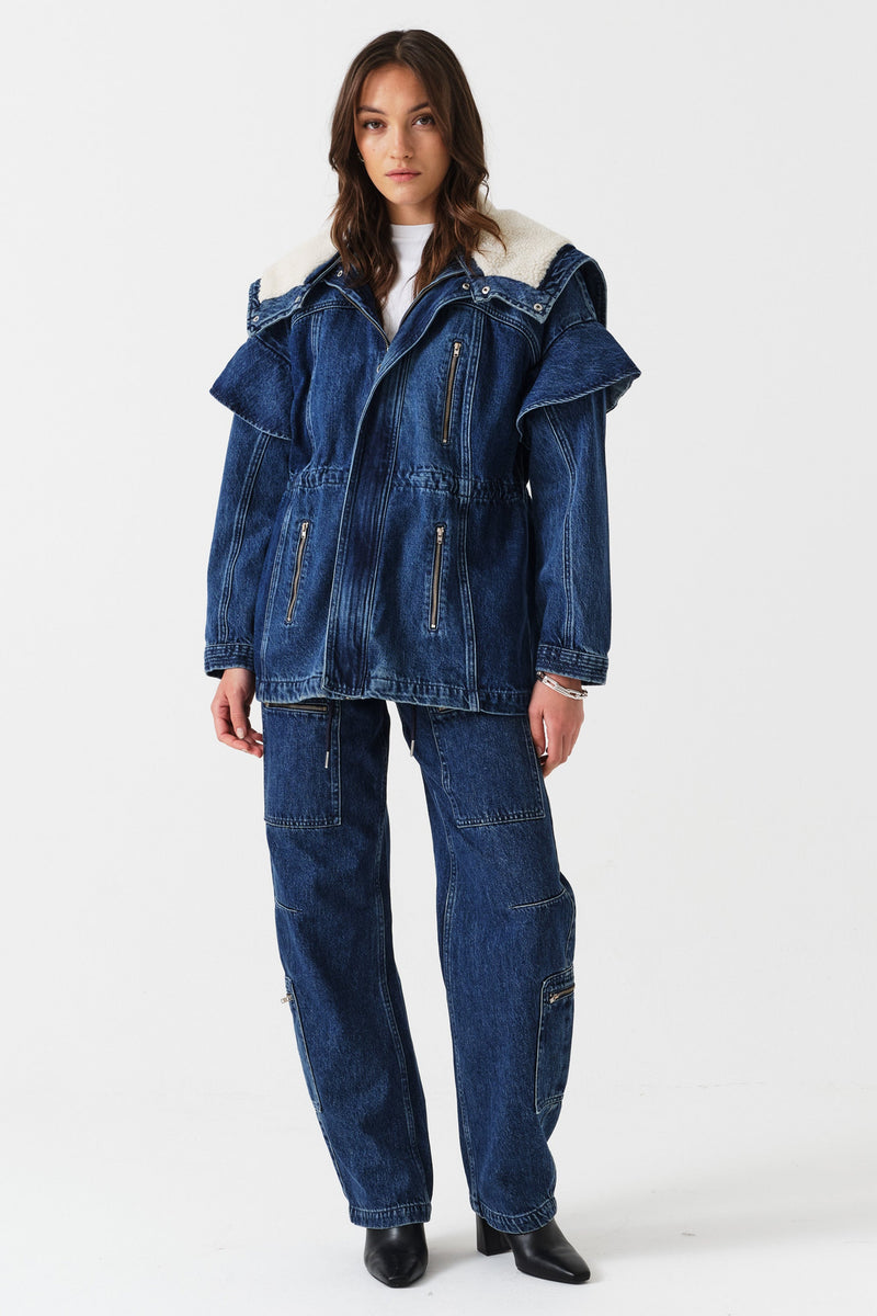 Woman wearing a white t-shirt, blue denim jacket with sherpa collar and zip details, and blue jeans with zip details on a white background 