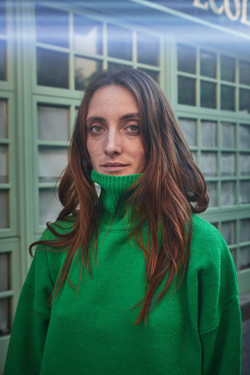 Woman wearing a green L'ENVERS sweater and light green skirt standing in front of a green door.