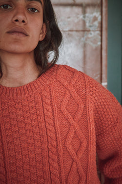 Person wearing an orange coral L'ENVERS sweater and white pants standing in a room with green doors and a marble wall.