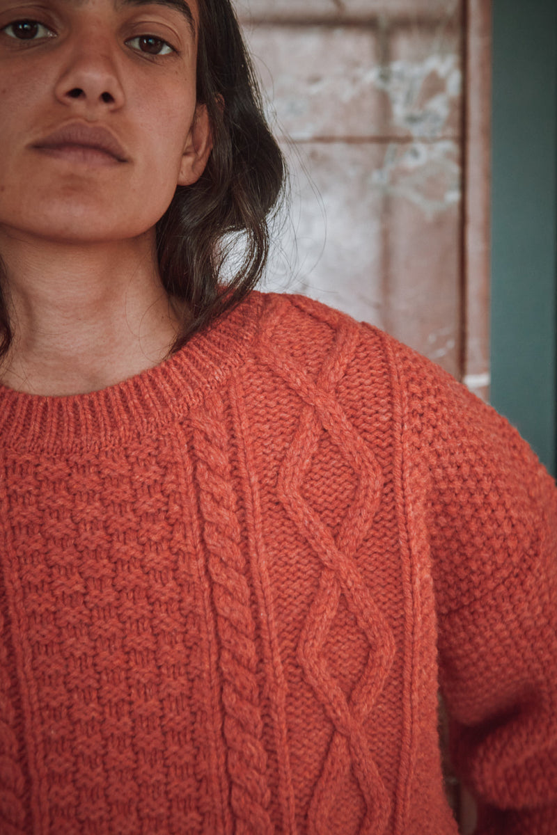 Person wearing an orange coral L'ENVERS sweater and white pants standing in a room with green doors and a marble wall.