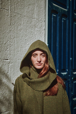 Person wearing a green hood with a matching pullover against a textured wall and blue door.