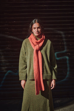 Person wearing a green pullover and coral scarf standing against a dark background