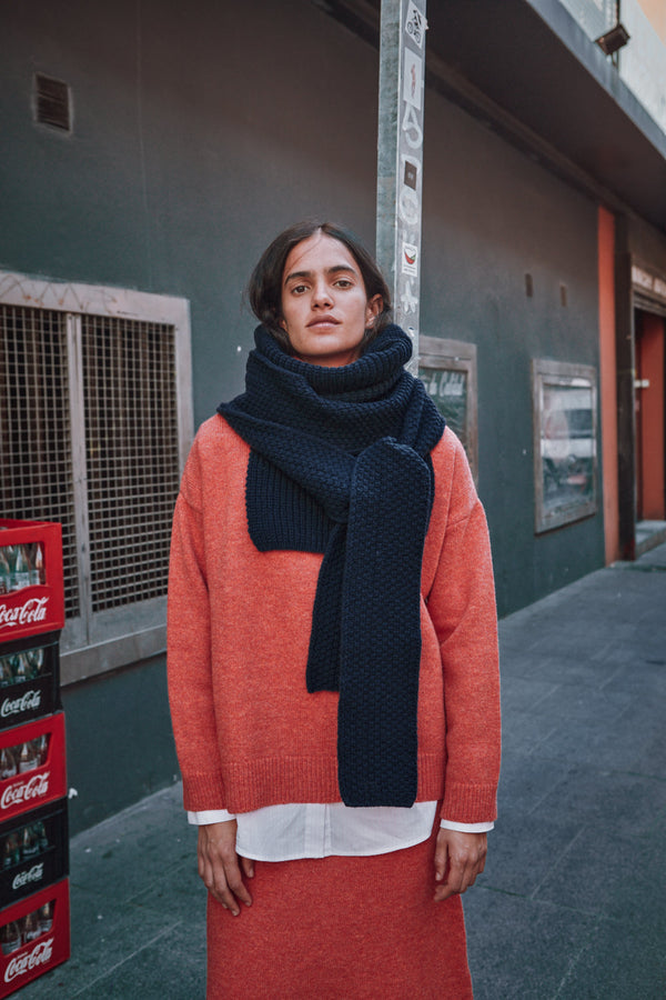 Person wearing an orange pullover and dark scarf standing next to Coca-Cola crates against a wall.