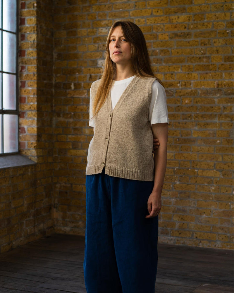 Woman wearing a beige vest and blue pants standing in a room with brick walls and wooden floor.