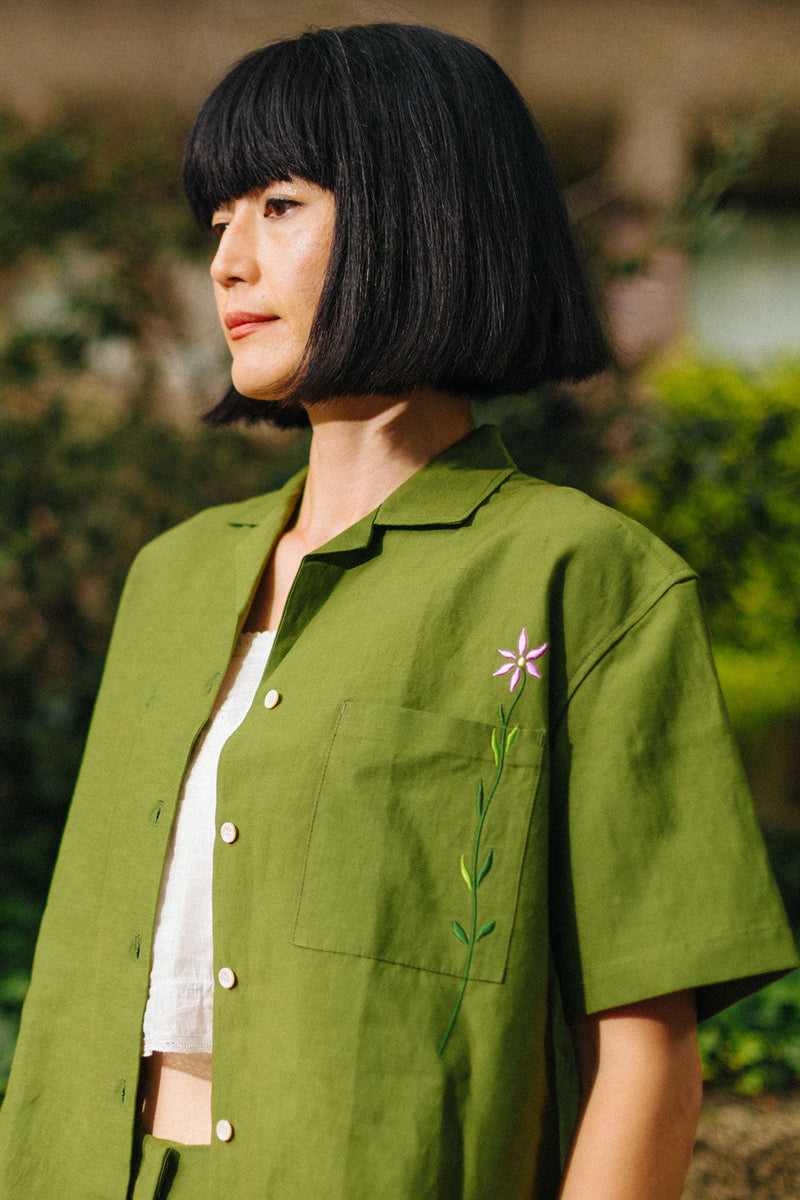 Woman wears Saywood's Frances Unisex Cuban Shirt in olive green. The image is a close up of the shirt, worn open over a white crop top. The green shirt is short sleeved, with an embroidered flower through the chest pocket. 