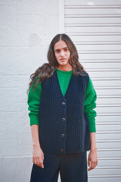 Woman wearing a green sweater and navy vest against a white wall.
