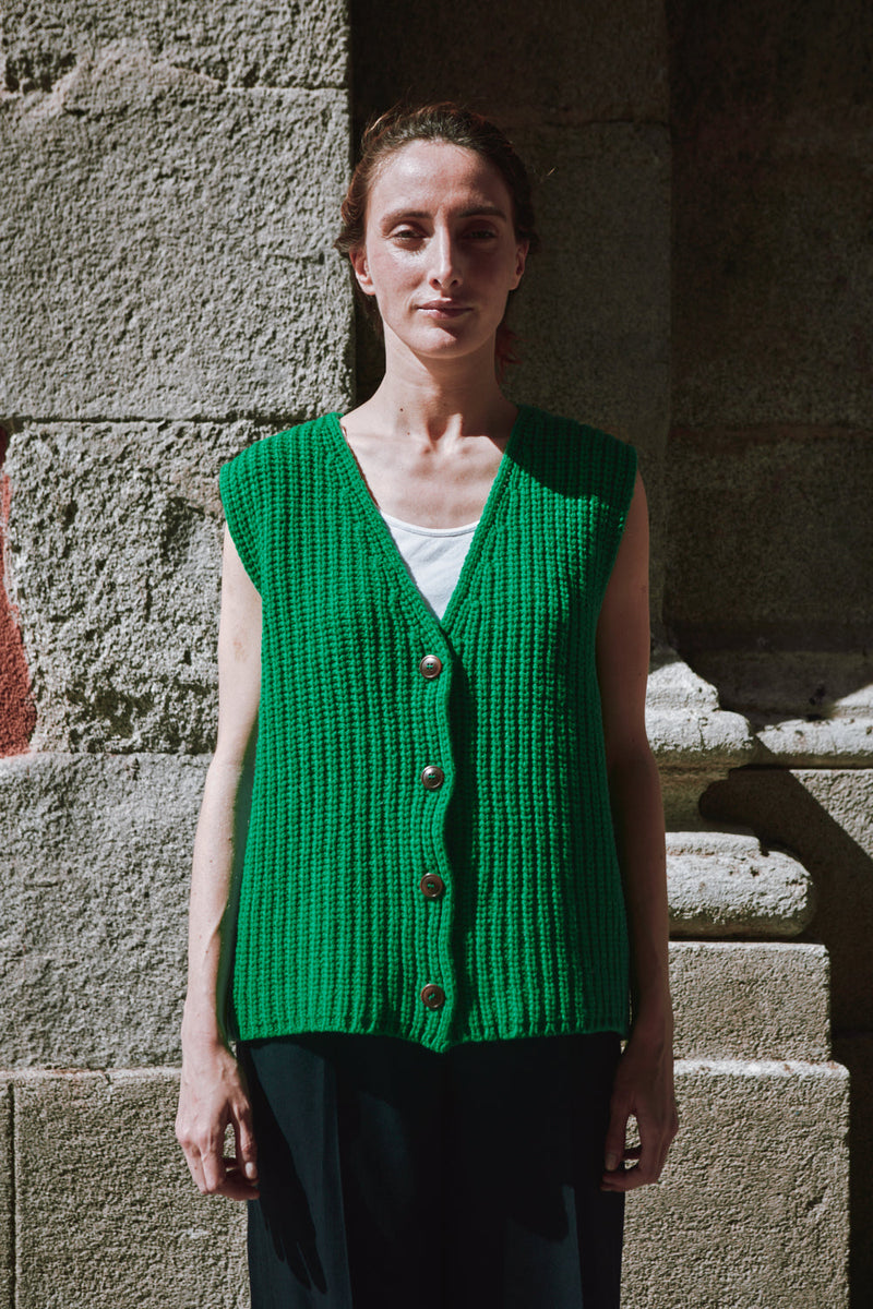 Person wearing a green knitted vest standing against a stone wall.