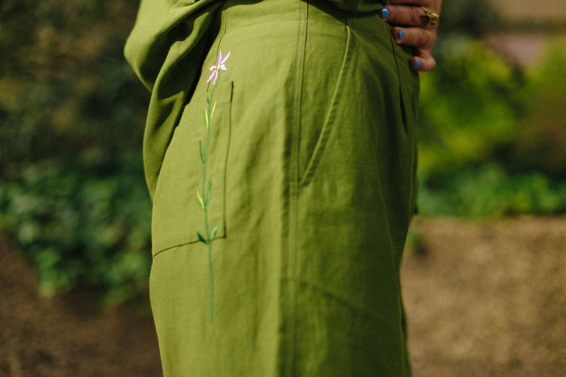 Close up of the side of the Bessie Shorts in olive green. The Saywood signature embroidered flower can be seen across the back patch pocket. The front angled side pocket on the shorts is visible, and greenery can be seen in the background.