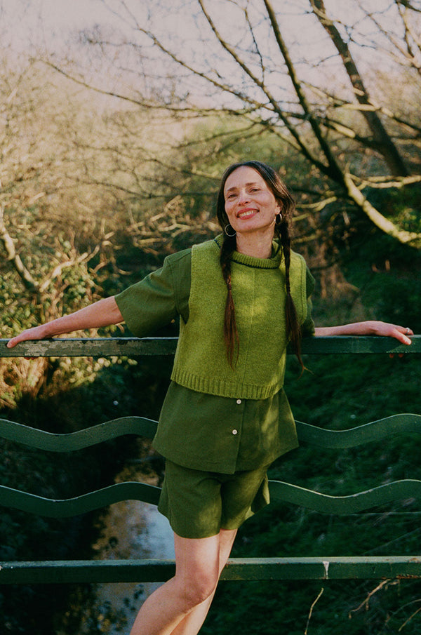A woman with plaited pigtail hair is standing in front of green railings on a bridge overlooking trees and a small river. She is leaning away from the rails and wearing Saywood's Bessie A-line Shorts in olive green, with matching Frances Unisex Cuban Short Sleeve Shirt. Worn with the new knitted wool sweater vest from Saywood.