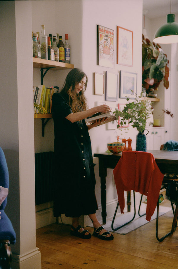 A woman stands in her kitchen diner reading a recipe book, leaning against a shelf of recipe books and drinks bottles, next to a French farmhouse style wooden table. She wears Saywood's Rosa Long Line Puff Sleeve Shirtdress in black Japanese denim, with black chunky sandals and with her hair worn down. There are flowers on the table, pictures on the all and a red cardigan is draped over the back of a chair.
