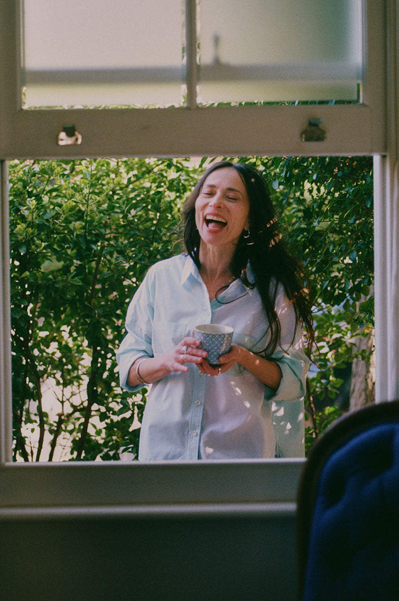 A woman is standing outside by tall green hedges, with the camera looking through an open sash window. She is holding a mug of tea and looking through the window, laughing. She is wearing Saywood's Lela Colourblock Boxy Shirt in green and blue pastel, with her sleeves rolled up.