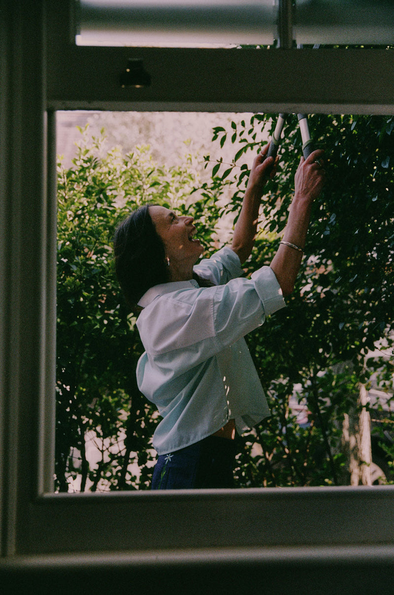 A woman is standing outside by tall green hedges, with the camera looking through an open sash window. She is holding hedge clippers and cutting the hedge. She is wearing Saywood's Lela Colourblock Boxy Shirt in green and blue pastel, with her sleeves rolled up. Worn with the Amelia Wide Leg Trousers in Japanese Denim. The signature embroidered flower can be seen on the back pocket of the trousers.