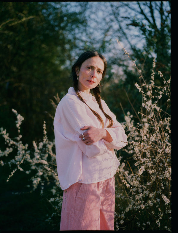 A woman stands in front of white flowering brambles, standing sideways to the camera, with her arms folded looking towards us. She wears Saywood's Amelia Wide Leg Trousers in pink corduroy with the Lela Colourblock Shirt in pink and orange. Her hair is in plaited pigtails.
