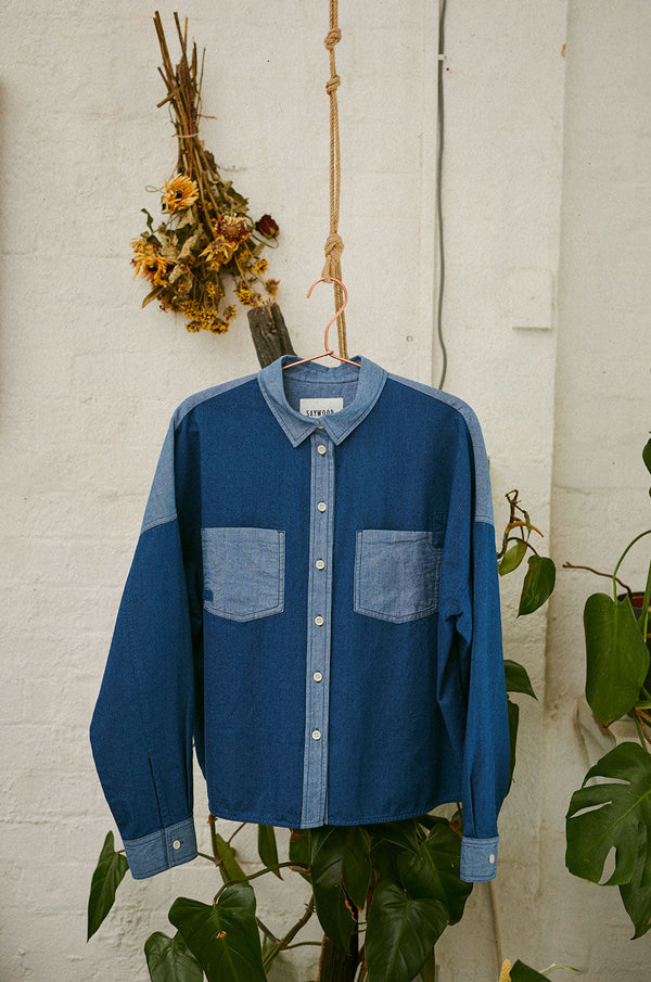 Saywood's Lela Patchwork Japanese Denim Shirt hangs on a rose gold hanger in front of a white brick wall on a rope. Behind it is a cheese plant and dried yellow flowers hanging on the wall.