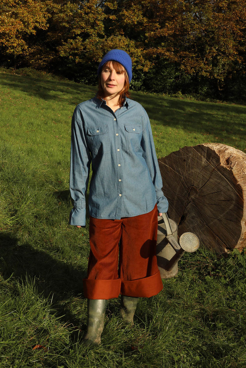 Women standing in a green field with trees behind her and a big tree trunk in the background. She is wearing Saywood's Caramel Tan Corduroy Trousers in Organic Cotton, styled rolled up with wellies, with the cotton Zadie Boyfriend Shirt in Japanese Denim, and a blue beanie hat. She holds a watering can.