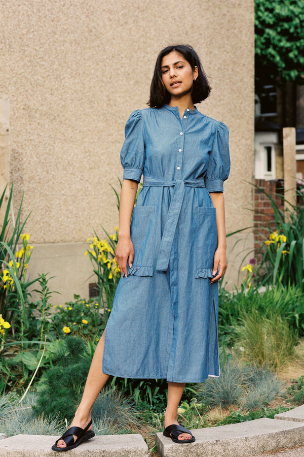 A woman stands wearing Saywood's Rosa denim shirtdress in light wash Japanese denim; a long line style with patch pockets and ruffles, a belt tied round the waist. She stands with her hands by her side at ease. Yellow flowers can be seen in the background.