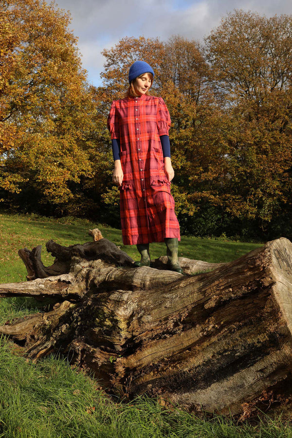 Woman stands in top of a fallen tree trunk in a field with trees and woodland behind her. She is wearing Saywood's Rosa Long Line Shirtdress in merlot and coral red tartan with a navy long sleeve top worn underneath and a blue beanie hat and green wellies.