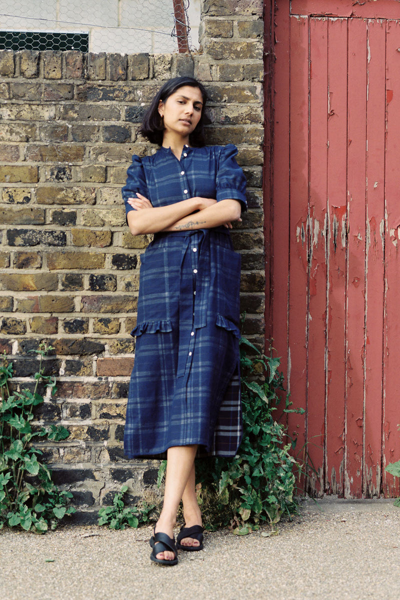 A woman leans against a brick wall with a red gate door next to her. She has her arms folded and wears Saywood's Rosa puff sleeve dress in navy check with black sandals. Plants can be seen growing out near her feet.