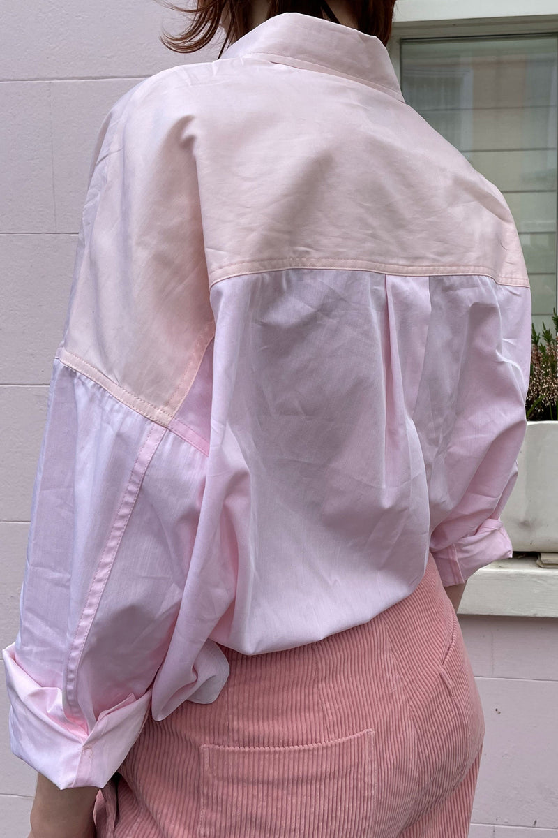 Woman stands in front of a pink brick house, wearing Saywood's boxy silhouette Lela Shirt in pastel pink and orange, tucked into a pair of pink corduroy trousers. Image shows the back of the shirt with the contrast back neck yoke and box pleat in the back body.